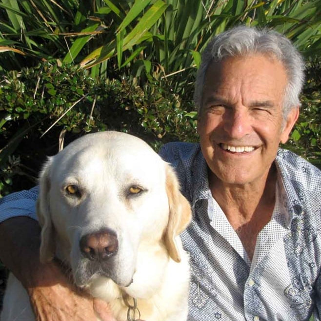 Smiling elderly man with gray hair sitting outdoors with large white Labrador dog near green plants