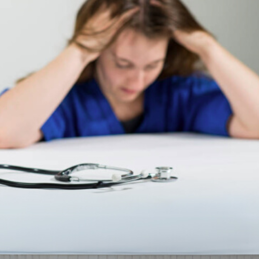 Stethoscope on table with stressed healthcare worker in blue scrubs holding their head in background