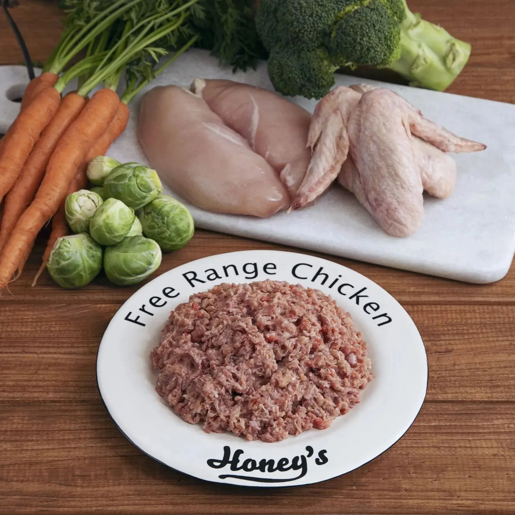 "Ground chicken patty on a plate with free range chicken, carrots, broccoli, and brussels sprouts on a wooden surface."