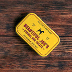 Yellow tin of Beautiful Joe's ethical dog treats on wooden surface with black dog silhouette
