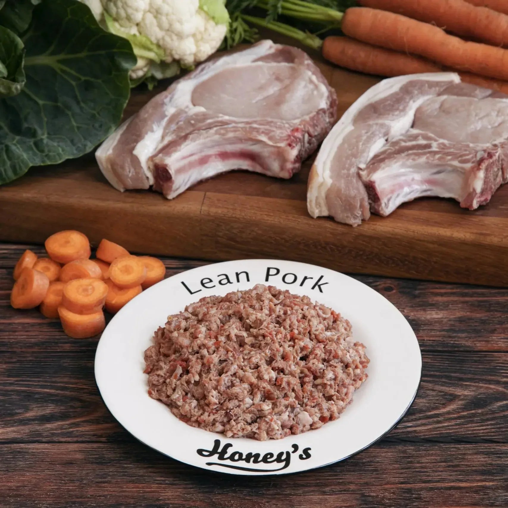 Plate of lean ground pork with raw pork chops, carrots, cauliflower, and greens on wooden surface